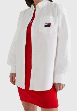 Kobieta ubrana w białą koszulę zapinaną na guziki z logo "Tommy Jeans" na kieszeni piersiowej, na ubranej czerwonej sukience.