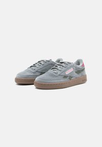 Tenisi gri din suede cu accente roz, sireturi plate, cutie pentru degete perforată și o talpă din cauciuc maro; au un logo Reebok pe lateral.
