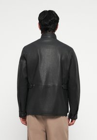 Black leather jacket with a high collar, textured surface, buttoned cuffs, and adjustable side straps. Simple and minimalistic design.