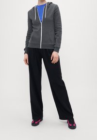 Sudadera gris con cremallera y capucha, bolsillos frontales, combinada con pantalones anchos negros y zapatos negros con acentos rosas. Camiseta azul visible.