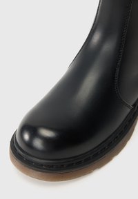 Bottine noire en cuir avec un bout arrondi, texture lisse et semelle en caoutchouc marron contrastante avec des détails de couture visibles.