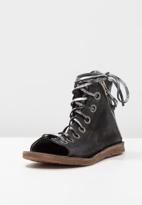 Black open-toe leather boot with silver laces, visible zipper on side, and brown worn sole on a white surface.