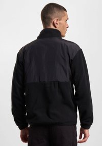 Svart fleece-jakke med en kontrasterende glatt svart panel over den øvre ryggen. Har høy krage og strikkede mansjetter.