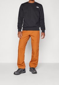 Felpa nera con colletto rotondo e logo bianco; pantaloni arancioni con tasche e cuciture; scarpe grigie con superficie testurizzata.