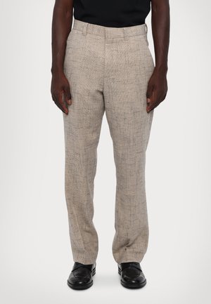 Pantalon beige texturé avec un motif subtil, coupe droite, passants de ceinture et poches latérales, associé à des chaussures noires.
