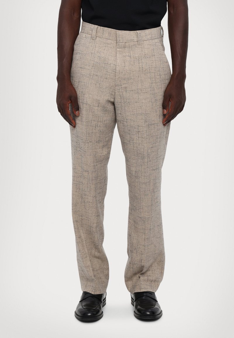 Beige textuurbroek met een subtiel patroon, rechte pijpen, riemlussen en zijzakken, gecombineerd met zwarte schoenen.