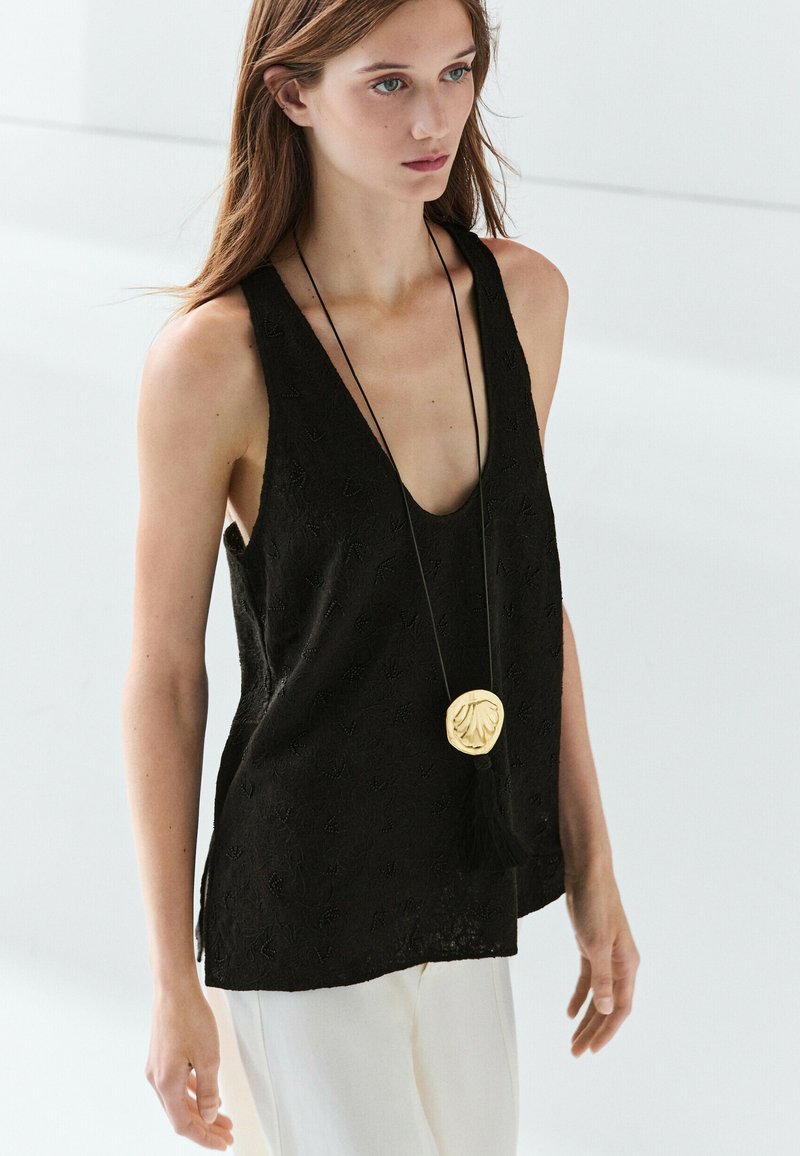 Young woman wearing a sleeveless black top with textured pattern, white pants, and a long necklace with a large gold pendant and black tassel.