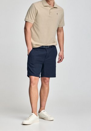 Man die een beige poloshirt, marineblauwe korte broek met riem en witte sneakers draagt, staand met één hand in de zak tegen een effen achtergrond.