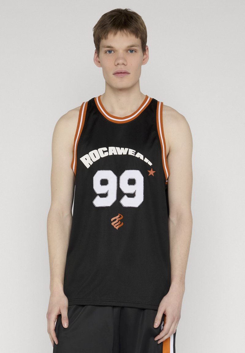 Jeune homme portant un maillot de basketball noir Rocawear avec le numéro 99 et une bordure orange et blanche, debout devant un fond uni.