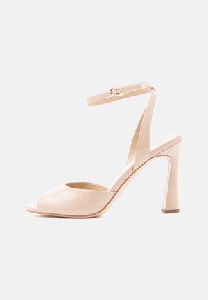 Sandale beige en cuir verni à talon haut avec bout ouvert, fine bride à la cheville, boucle dorée et talon bloc incurvé sur fond blanc.