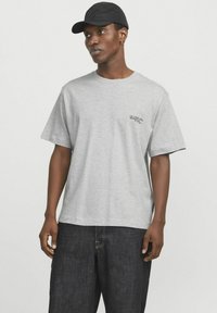 Camiseta de algodón gris de manga corta, con un pequeño logotipo impreso en negro en el lado izquierdo. Combinada con pantalones de mezclilla oscuros.