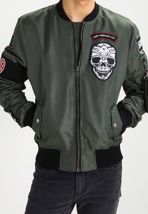 Veste bomber vert olive avec poignets noirs, écusson tête de mort avec un œil, et texte "Light motorcycle club" sur la poitrine. Mains dans les poches.
