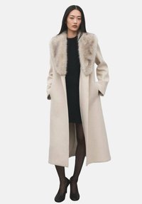 Abrigo largo beige con cuello de piel sintética, corte entallado, bolsillos laterales y una silueta limpia, llevado sobre un vestido negro y medias.