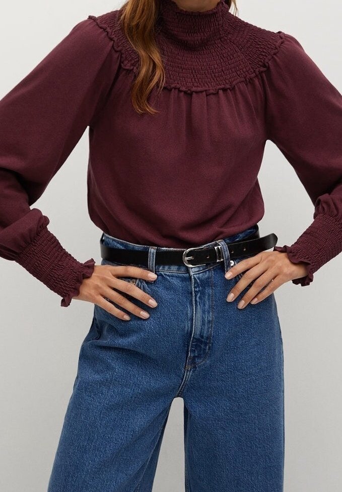 Bordeaux lange mouw blouse met hoge smock-kraag en geplooide mouwen, gecombineerd met hoog opgesneden blauwe denim jeans en een zwarte riem.