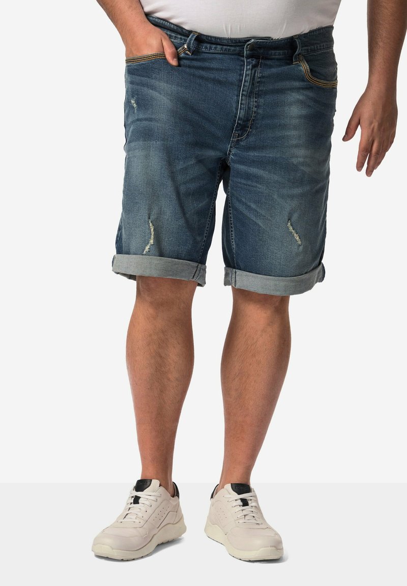 Denim šortky v středně modrém odstínu s ohrnutými lemy, s opotřebovanou texturou a viditelnými švy. Spárované s bílými teniskami.