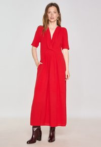 Red maxi dress with a deep V-neck and short sleeves, featuring a knotted bodice detail and side pockets, paired with heeled brown boots.