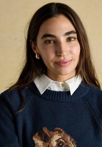 Tumes sinise kudumi aluskarp, millel on detailne looma tikand, kantud valge kaelusega särk üle. Kuldsed rõngaskõrvarõngad täiendavad välimust.