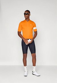 Kortærmet orange cykeltrøje med lynlås foran. Sorte polstrede shorts, hvide sokker og sneakers. Holder en vandflaske og har solbriller på.