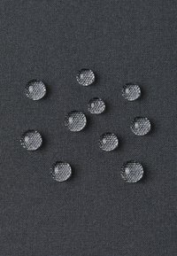 Gocce d'acqua limpida su un tessuto scuro e strutturato, di varie dimensioni, che mostrano una forma arrotondata e una superficie liscia, riflettendo la luce.