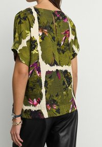 Zielona kwiatowa bluzka z krótkimi rękawami, z białymi abstrakcyjnymi liniami, z wycięciem na plecach i teksturowanym materiałem. Połączona z czarnymi spodniami.