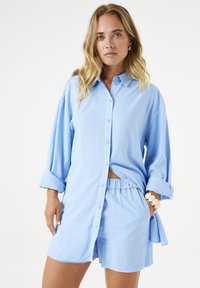 Camisa de botones azul claro con mangas largas y enrolladas, combinada con pantalones cortos a juego. Tejido suave con un corte holgado y cinturilla elástica.