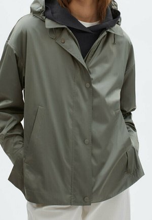 Personne portant une veste à capuche vert olive clair par-dessus un sweat à capuche noir, les mains dans les poches de la veste, visible des épaules aux hanches.