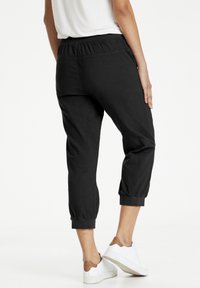 Henkilö, jolla on päällään mustat lyhyet jogger-housut ja valkoiset lenkkarit, seisoo yksi käsi lantiolla tasaisella valkoisella taustalla.