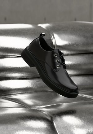 Chaussure noire en cuir lisse à lacets suspendue au-dessus d'une surface tuftée métallique argentée brillante, mettant en valeur ses détails polis.