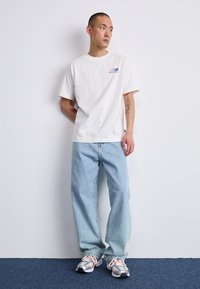 Un tricou alb din bumbac cu un mic logo multicolor, asociat cu blugi de denim oversized de culoare albastru deschis și adidași gri cu accente portocalii.