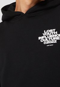 Sweat à capuche noir avec texte blanc sur la poitrine indiquant « LOST YOUTH INFLUENCE CULTURE ©2022 », porté par une personne visible de la nuque jusqu'en bas.