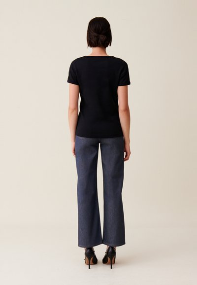 T-shirt noir à manches courtes associé à un pantalon flare en denim foncé taille haute et des chaussures à talons noires, vue de dos. Texture de tissu lisse.