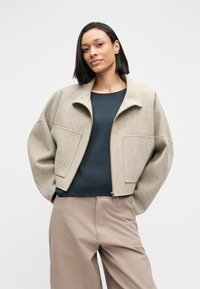 Beżowa kurtka z mieszanki wełny z zamkiem z przodu, krótkim fasonem, dużymi naszywanymi kieszeniami i wysokim kołnierzem, zestawiona z granatową bluzką i beżowymi spodniami.