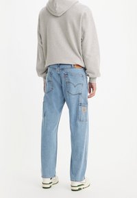 Person som bär ljusblå denimjeans med bak- och sidofickor, en ljusgrå hoodie och vita sneakers, stående mot en vit bakgrund.