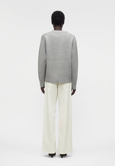 Άτομο με κοντά σγουρά μαλλιά που φορά γκρι πουλόβερ με μακριά μανίκια και λευκό παντελόνι με φαρδιά πόδια, στέκεται μπροστά σε απλό φόντο.