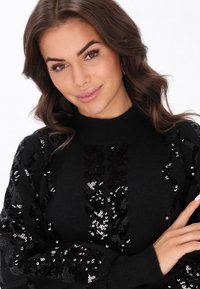 Fekete kötött pulóver, magas gallérral, textúrált levélmintával díszítve, flitterekkel az ujjakon és az elején.