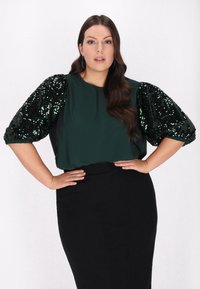 Blusa verde con maniche a sbuffo tempestata di paillettes, abbinata a una gonna nera a tubino. Tessuto morbido, dettagli lucidi e vestibilità rilassata.