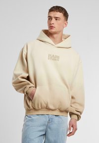 Beige huvtröja med en framficka och ribbade muddar. "KANI STUDIOS"-logotyp i ljusare beige syns på bröstet. Strukturerad tyg.