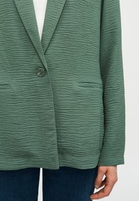 Πράσινο σακάκι με υφή με κλείσιμο με ένα κουμπί, φορεμένο πάνω από λευκό τοπ και σκούρο μπλε τζιν, με το χέρι χαλαρό στο πλάι.