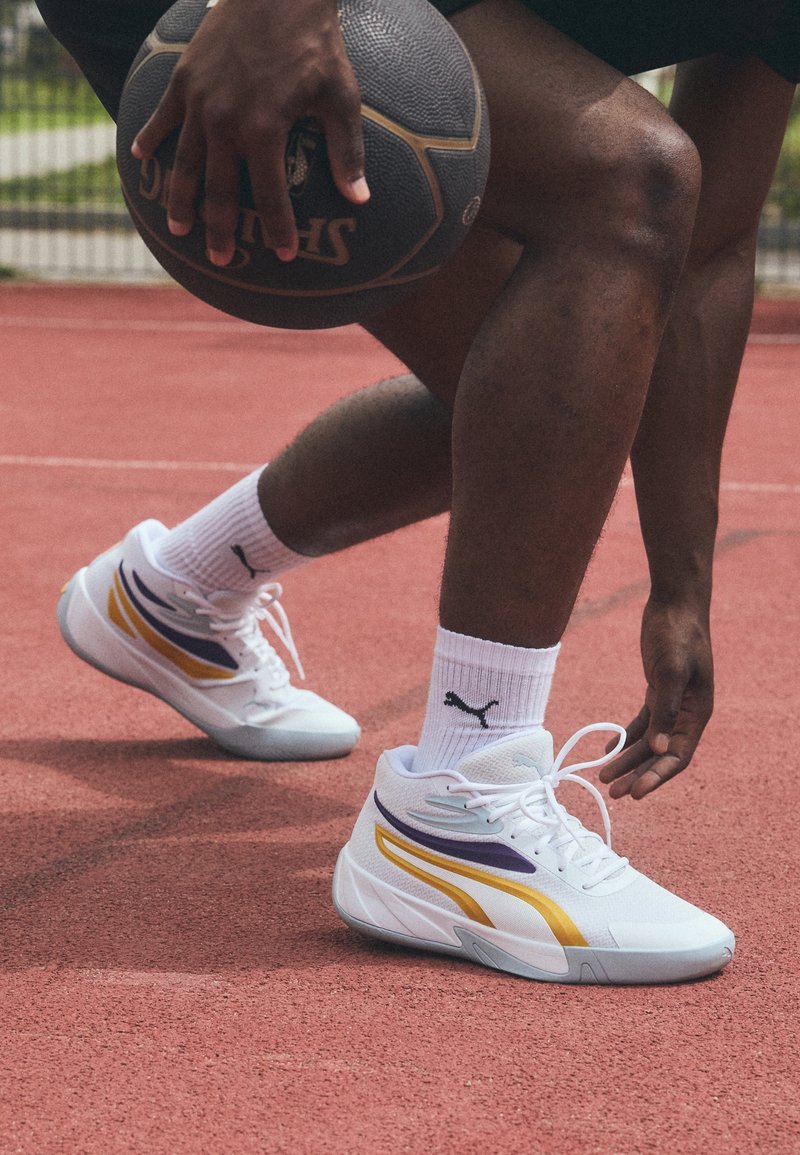 Baskets montantes blanches avec des accents violets et dorés, dessus en mesh texturé et semelle en caoutchouc. Un athlète est accroupi sur un terrain de basket.