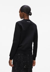 Musthave mustam svart ülikond, millel on ümmargune kaelus ja soonikkoes käised. Seljal on Karl Lagerfeldi logo valge värviga. Kanded koos sädelevate mustade seelikutega.