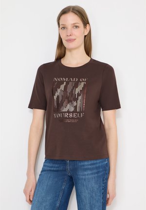 Kobieta w ciemnobrązowej koszulce z napisem "Nomad of Yourself" i abstrakcyjnym wzorem, w niebieskich dżinsach, stojąca na białym tle.