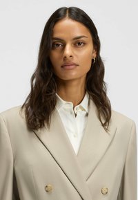 Beige double-breasted blazer with a notched lapel and two large buttons, layered over a white collared shirt. Smooth fabric texture.