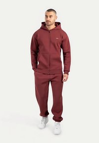 Sudadera con cremallera burdeos y pantalones a juego hechos de tela suave y gruesa. Cuenta con capucha, bolsillos delanteros y puños acanalados. Se combina con zapatillas blancas.