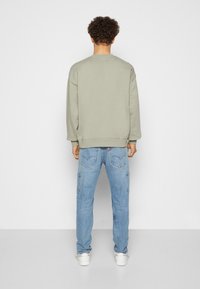 Sudadera verde con puños y dobladillo acanalados, combinada con vaqueros cargo azul claro con bolsillos laterales y un logo en la parte trasera. Zapatillas blancas.