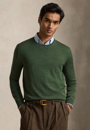 Homme portant un pull vert à col rond sur une chemise bleue à col, pantalon marron à carreaux et ceinture, debout les mains dans les poches.