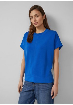 Frau mit braunen Haaren, die ein hellblaues kurzärmeliges Oberteil und blaue Jeans trägt, steht vor einer schlichten hellgrauen Wand.