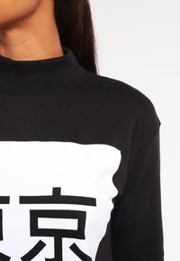 Musta poolo, jossa on suuri valkoinen suorakulmainen grafiikka mustalla tekstillä. Kangas vaikuttaa pehmeältä ja sileältä.