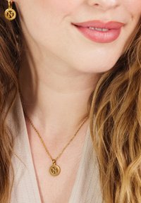 Gouden ketting met een ronde hanger met een gegraveerd "N"-ontwerp, hangend aan een delicate ketting. Achtergrond van textuur op huid en haar.