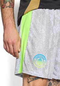 Grå sportsbukser med et tekstureret vanddråbemønster og neongrøn sidelæaf. Har et Manchester City-logo på sømmen.
