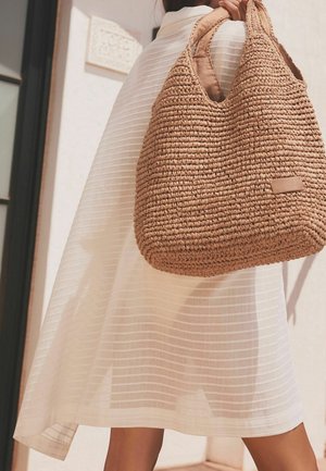 Grote tan geweven tas met een gestructureerd ontwerp, voorzien van twee handvatten en een klein leren label. De achtergrond bevat een gedrapeerde wit gestreepte stof.